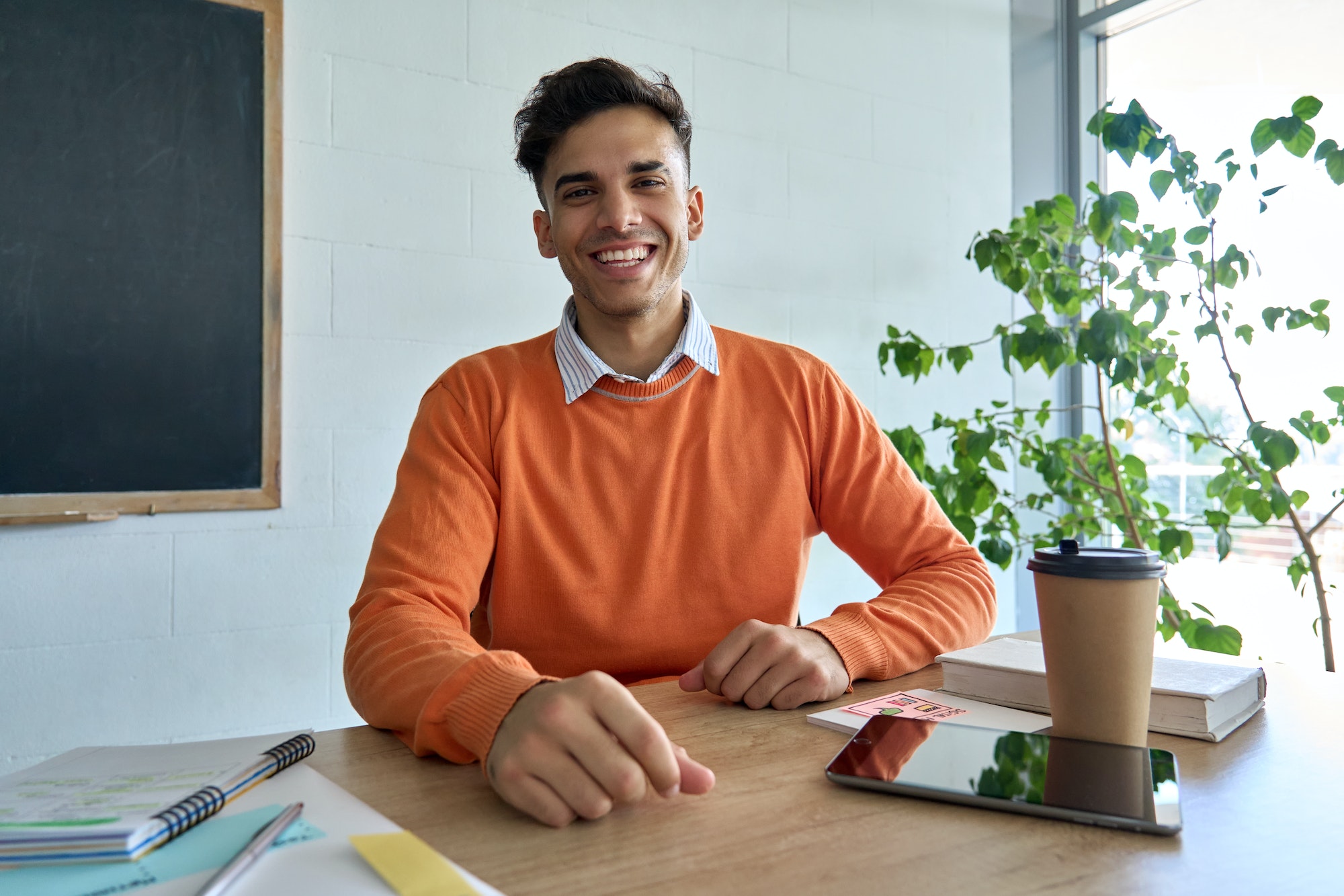 Happy smiling Indian student looking at camera sitting at desk in classroom.